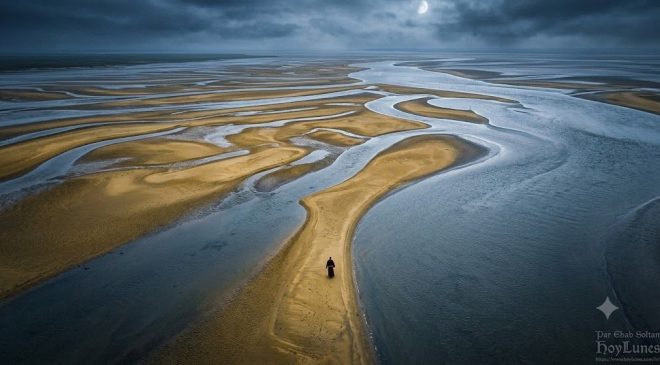 Une vue aérienne spectaculaire (plan zénithal large) montrant une silhouette minuscule vêtue d'une robe de moine traversant les langues de sable infinies et labyrinthiques de la baie. L'eau de la marée montante commence à cerner les chemins sablonneux, créant des courbes organiques argentées sur la terre. L'immensité du paysage souligne la vulnérabilité absolue du marcheur.
