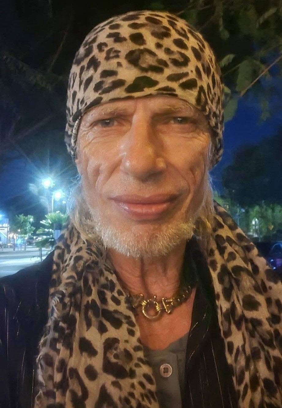 Un primer plano de Frankie Ray sonriendo, con un pañuelo de leopardo en la cabeza y una bufanda a juego, de noche.