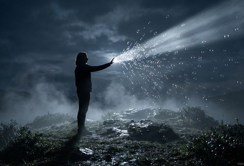 Una figura humana de perfil sobre la cima de una colina nocturna rodeada de niebla baja. La persona extiende la mano intentando atrapar un haz de luz de luna denso y plateado que atraviesa la oscuridad. En el punto exacto donde la mano toca la luz, esta se fragmenta en diminutas burbujas de jabón luminosas y efímeras que se disuelven en el aire.
